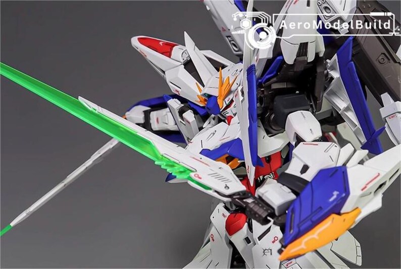 Puede incluir: Modelo detallado de un robot mecha blanco, azul, rojo y amarillo con una espada de energ&iacute;a verde. El robot tiene dise&ntilde;os intrincados y est&aacute; posado din&aacute;micamente. El texto "AeroModelBuild" es visible en la esquina superior derecha.