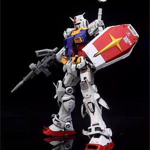 Op de afbeelding: Gedetailleerd model van een Gundam-robot, voornamelijk wit met rode, blauwe, gele en grijze accenten. Het houdt een grijs geweer en een rood schild met een gele ster vast. Het model staat tegen een zwarte achtergrond.
