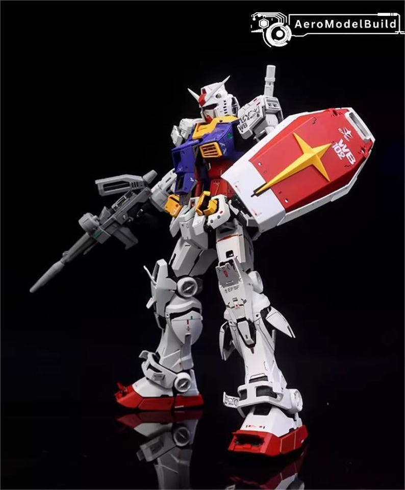 Op de afbeelding: Gedetailleerd model van een Gundam-robot, voornamelijk wit met rode, blauwe, gele en grijze accenten. Het houdt een grijs geweer en een rood schild met een gele ster vast. Het model staat tegen een zwarte achtergrond.