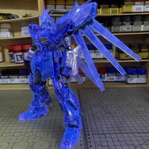 May include: A transparent blue model robot with articulated limbs and large wing-like structures. The model is posed on a cutting mat with a ruler, and is set against a backdrop of shelves with paint and tools.