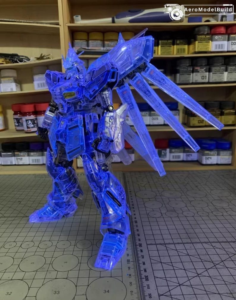 May include: A transparent blue model robot with articulated limbs and large wing-like structures. The model is posed on a cutting mat with a ruler, and is set against a backdrop of shelves with paint and tools.