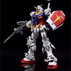 Op de afbeelding: Gedetailleerd model van een Gundam-robot, voornamelijk wit met rode, blauwe, gele en grijze accenten. Het houdt een grijs geweer en een rood-wit schild met een gele ster vast. Het model staat op een reflecterend zwart oppervlak.
