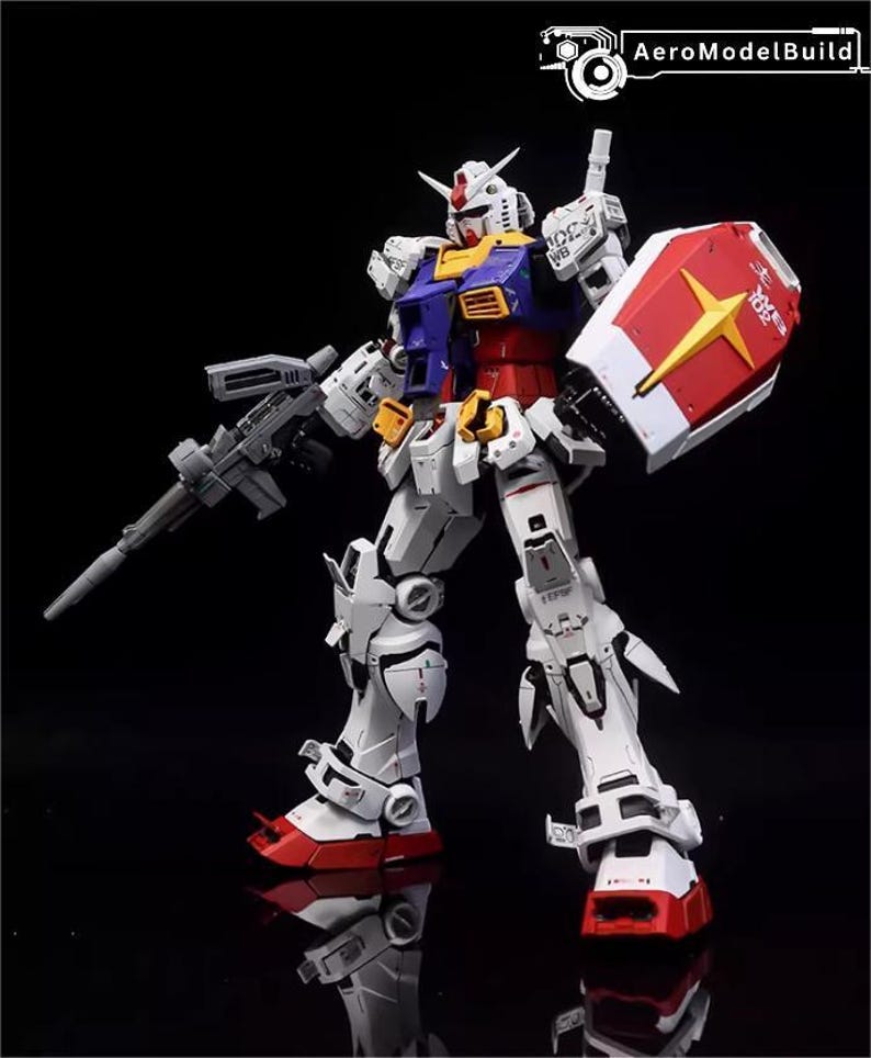 Op de afbeelding: Gedetailleerd model van een Gundam-robot, voornamelijk wit met rode, blauwe, gele en grijze accenten. Het houdt een grijs geweer en een rood-wit schild met een gele ster vast. Het model staat op een reflecterend zwart oppervlak.