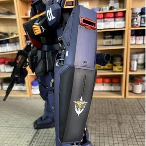 Pu&ograve; includere: Modello dettagliato di un robot mecha blu scuro e nero con accenti arancioni e rossi. Il robot ha uno scudo con un logo e il numero "01" sulla spalla. Lo sfondo include scaffali con varie bottiglie e contenitori.