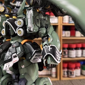 May include: Close-up of a detailed, olive green and black model robot with gold accents. The robot has intricate mechanical parts, including panels, joints, and circular components. The text "AeroModelBuild" is visible in the upper right corner.