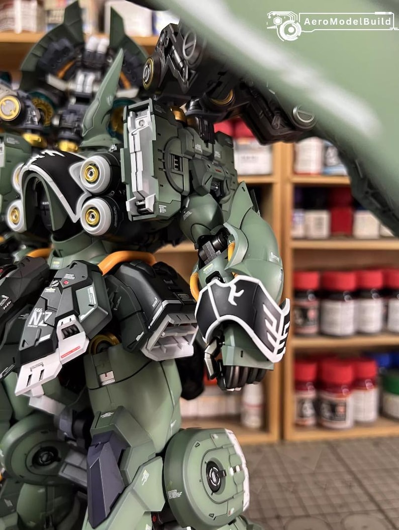 May include: Close-up of a detailed, olive green and black model robot with gold accents. The robot has intricate mechanical parts, including panels, joints, and circular components. The text "AeroModelBuild" is visible in the upper right corner.