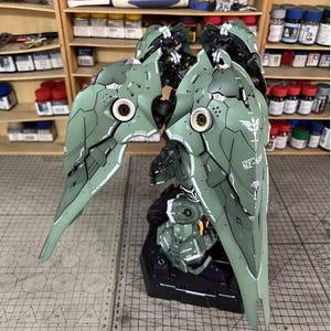 May include: Assembled model of a green and white mecha with detailed panel lines and markings. The model features large, wing-like structures with intricate designs and the text "NZ-66". The model is on a black base, with a cutting mat in the foreground.