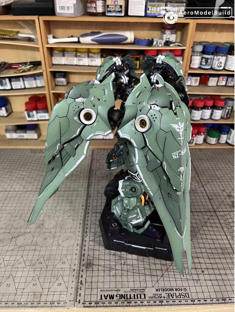 May include: Assembled model of a green and white mecha with detailed panel lines and markings. The model features large, wing-like structures with intricate designs and the text "NZ-66". The model is on a black base, with a cutting mat in the foreground.