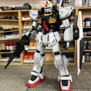 May include: A detailed model of a Gundam robot, primarily white with black, red, and yellow accents. It holds a black rifle and a white shield with yellow and black details. The model is on a cutting mat, with the text "ArrowModelBuild" visible.