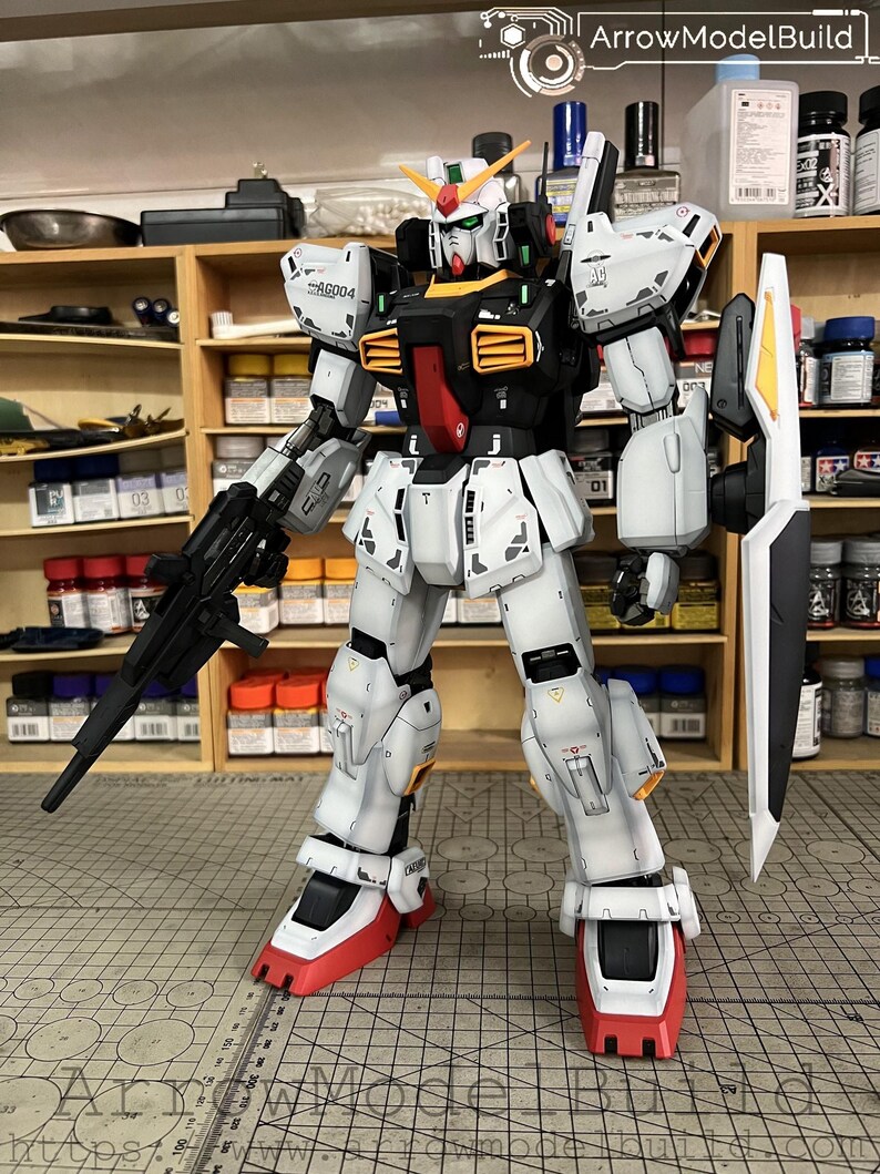 May include: A detailed model of a Gundam robot, primarily white with black, red, and yellow accents. It holds a black rifle and a white shield with yellow and black details. The model is on a cutting mat, with the text "ArrowModelBuild" visible.