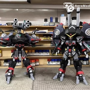 May include: Two detailed model robots, one with multiple cannons and the other with large circular shoulder armor. The robots are primarily black, red, and gray, with intricate designs and details. The background shows shelves with various tools and supplies.