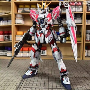 May include: A detailed model of a Gundam robot, primarily white with red and pink accents. The robot is holding a rifle and a large shield. The model is standing on a surface with a ruler.