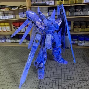May include: A clear blue model robot kit, featuring detailed design and sharp edges. The robot has large wings and is positioned on a cutting mat. The background shows shelves with paint jars and model supplies.