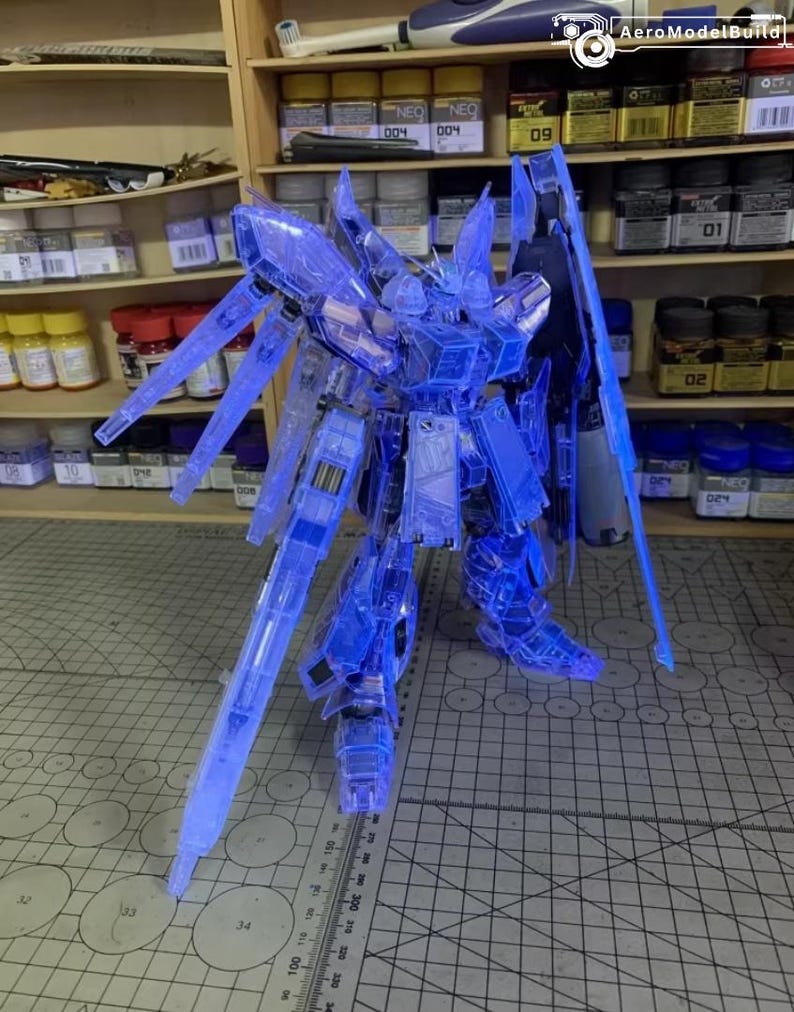 May include: A clear blue model robot kit, featuring detailed design and sharp edges. The robot has large wings and is positioned on a cutting mat. The background shows shelves with paint jars and model supplies.