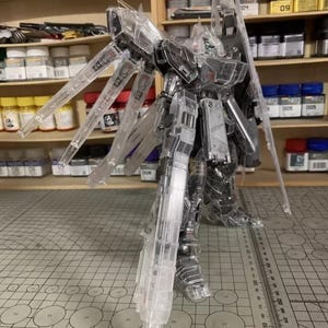May include: A clear, transparent model kit of a robot with articulated limbs and wings. The model is standing on a cutting mat with a ruler. The background includes shelves with paint bottles and the AeroModelBuild logo.