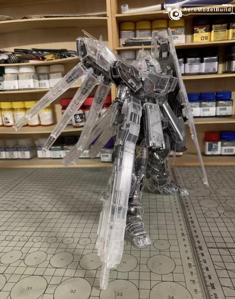 May include: A clear, transparent model kit of a robot with articulated limbs and wings. The model is standing on a cutting mat with a ruler. The background includes shelves with paint bottles and the AeroModelBuild logo.
