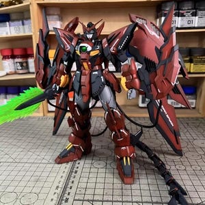 May include: A detailed model of a Gundam robot, primarily in shades of red, black, and gray. The robot has intricate armor, large wings, and a green energy sword. The model is posed on a cutting mat, showcasing its design and craftsmanship.