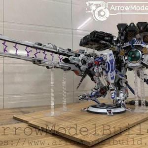 May include: A detailed model of a futuristic robot with a large cannon and multiple attachments. The robot is primarily white, blue, and gray, with purple lightning bolt accents. The model is displayed on a wooden base with the text "ArrowModelBuild".