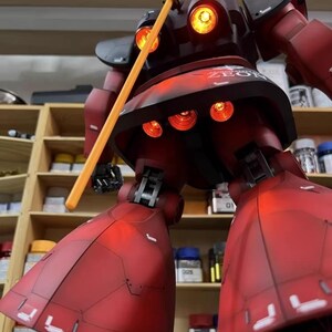 May include: A detailed model of a red and black robot with illuminated orange lights. The robot is holding a yellow staff and features the text "ZEON". The model is displayed in front of a shelf with various containers.
