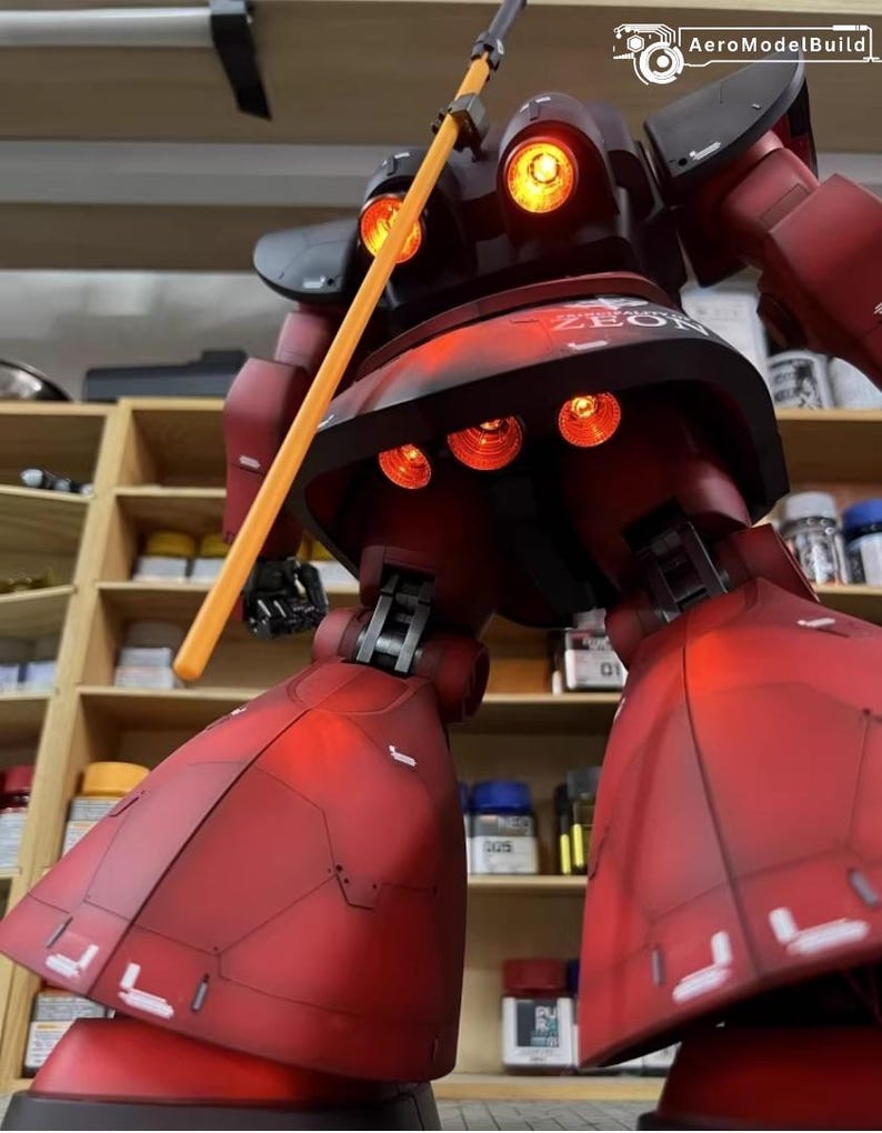 May include: A detailed model of a red and black robot with illuminated orange lights. The robot is holding a yellow staff and features the text "ZEON". The model is displayed in front of a shelf with various containers.