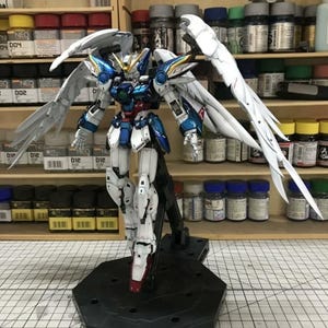 May include: A Wing Gundam Zero model kit featuring a metallic blue and white color scheme with large articulated white wings, a yellow V-fin, and a green chest sensor, mounted on a black hexagonal display stand on a grid cutting mat.