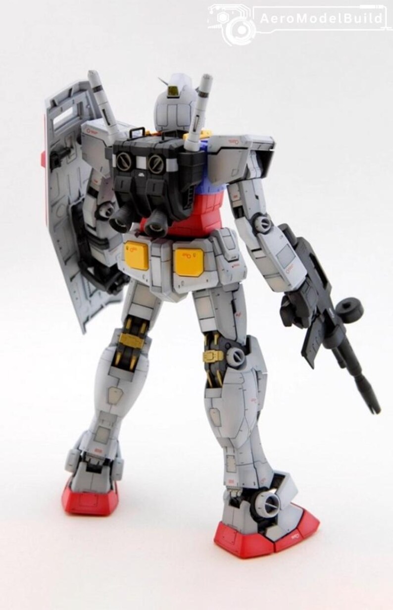 May include: A detailed model of a Gundam robot, primarily gray with red, yellow, and blue accents. It holds a black rifle and a shield. The model is standing with its legs apart, showcasing its mechanical design.