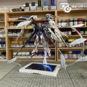 May include: A Wing Gundam Zero model kit featuring a blue, white, and red color scheme with large articulated wings. The figure is mounted on a white display base with a blue graphic, surrounded by a circular arrangement of white feather-like parts.