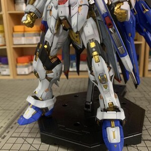 Puede incluir: Un kit de modelo detallado de un robot mecha, con una combinaci&oacute;n de colores blanco, dorado, azul y negro. El robot est&aacute; posado sobre un soporte negro, mostrando intrincados detalles mec&aacute;nicos y l&iacute;neas de panel. El texto "ArrowModelBuild" es visible.