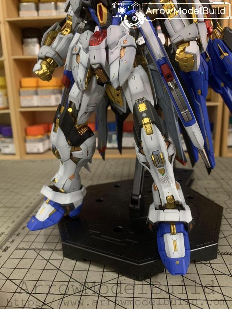 Puede incluir: Un kit de modelo detallado de un robot mecha, con una combinaci&oacute;n de colores blanco, dorado, azul y negro. El robot est&aacute; posado sobre un soporte negro, mostrando intrincados detalles mec&aacute;nicos y l&iacute;neas de panel. El texto "ArrowModelBuild" es visible.