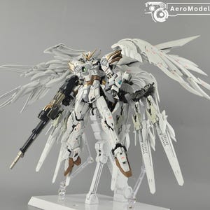 May include: A white and gold model robot with large, detailed wings and a long gun. The robot is on a white stand against a gray background. The text "AeroModelBuild" is in the upper right corner.