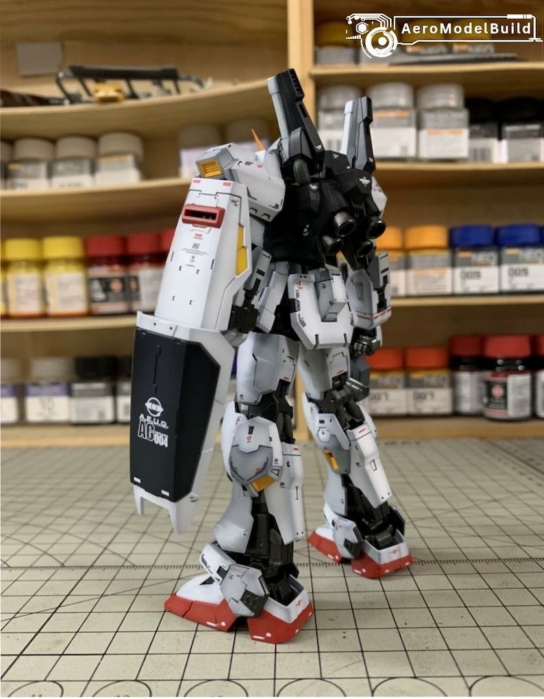 Pode incluir: Modelo detalhado de um rob&ocirc; branco, preto e cinza com detalhes em vermelho. O rob&ocirc; tem um escudo com o texto "A.E.U.G. AC004" e est&aacute; posicionado sobre uma superf&iacute;cie quadriculada. Provavelmente &eacute; um item de colecionador ou um projeto de hobby.