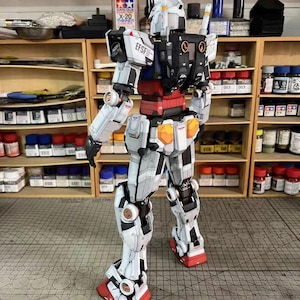 May include: A detailed, assembled Gundam model kit, predominantly white with black, red, and orange accents. The model features intricate mechanical details, visible joints, and the text "EFSF". The model is posed on a cutting mat.