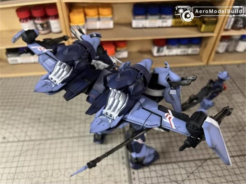 May include: A detailed model of a futuristic aircraft, primarily in shades of blue and gray, with intricate mechanical details. The model features wings, a cockpit, and various weapon-like attachments. The background includes shelves with paint bottles.