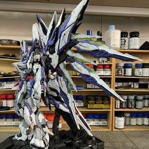 May include: A detailed model of a futuristic robot with white, blue, and purple armour. The model has large, stylized wings and is mounted on a black base. The robot is posed in a dynamic stance, showcasing intricate details.