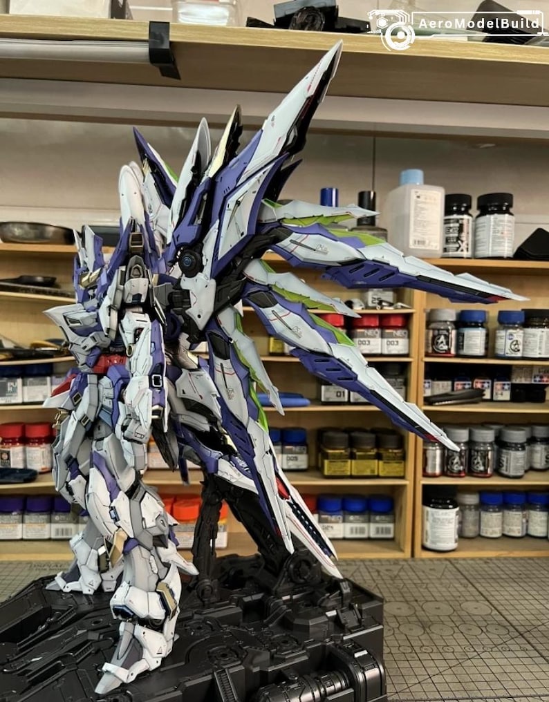 May include: A detailed model of a futuristic robot with white, blue, and purple armour. The model has large, stylized wings and is mounted on a black base. The robot is posed in a dynamic stance, showcasing intricate details.