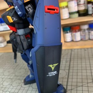 May include: A detailed model of a mecha robot, primarily in shades of blue, black, and yellow. The robot features a large shield with the text "TITANS GUNDAM PROJECT RX-178" and a stylized eagle emblem. The model is highly detailed with visible mechanical components.