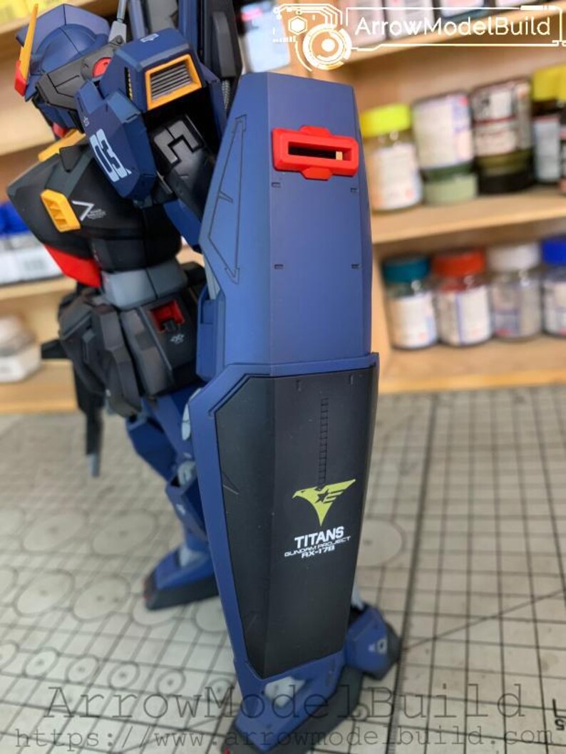 May include: A detailed model of a mecha robot, primarily in shades of blue, black, and yellow. The robot features a large shield with the text "TITANS GUNDAM PROJECT RX-178" and a stylized eagle emblem. The model is highly detailed with visible mechanical components.