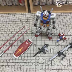 以下が含まれることがあります： 白、青、赤、黄色の配色が特徴のガンダムロボットの精密模型キット。 シールド、武器、長い赤い槍などの様々なアクセサリーが含まれています。 モデルはグリッドパターンの表面に配置されています。