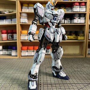 May include: A detailed, white and gray model robot with blue accents, standing on a grid surface. The robot has intricate armor plating and is posed with one leg forward. The background includes shelves with paint bottles and the text "ArrowModelBuild".