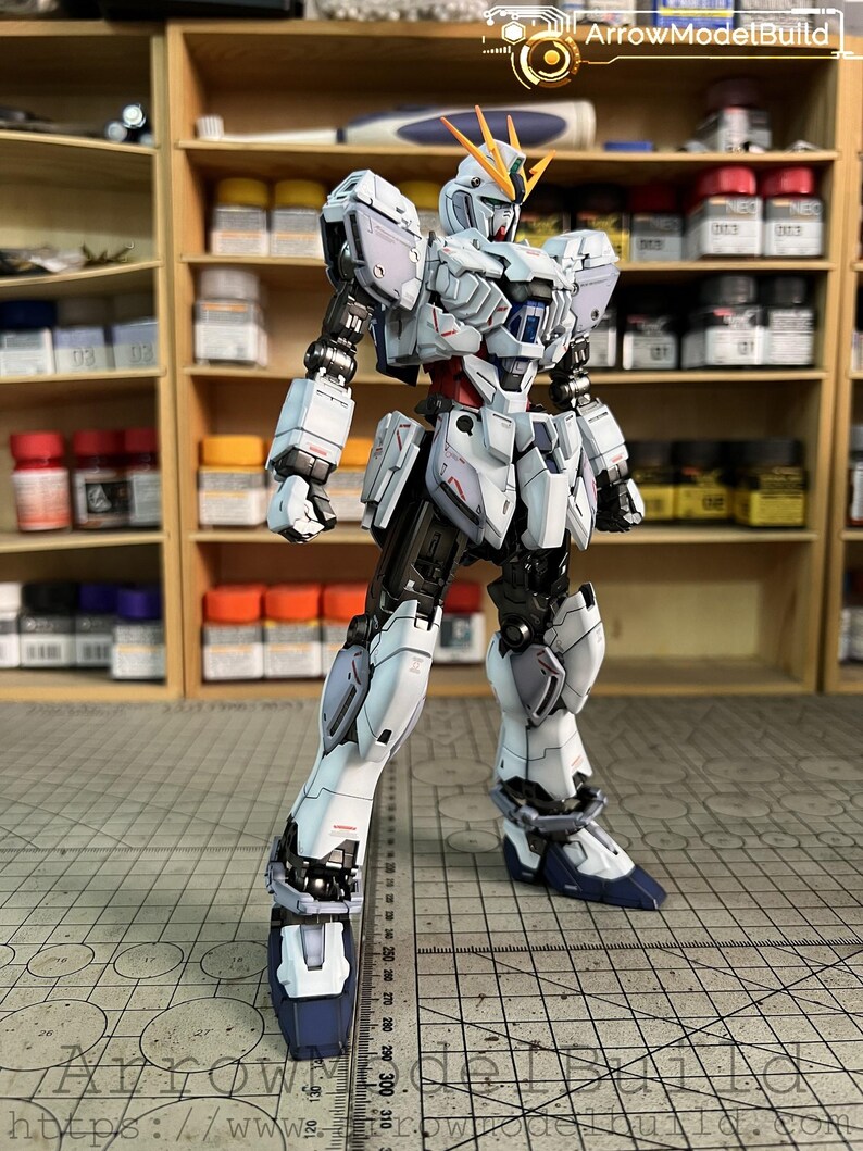 May include: A detailed, white and gray model robot with blue accents, standing on a grid surface. The robot has intricate armor plating and is posed with one leg forward. The background includes shelves with paint bottles and the text "ArrowModelBuild".