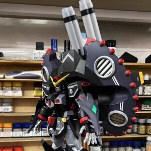 May include: A detailed model of a robot with a black, gray, and red color scheme. The robot has large shoulder-mounted cannons and intricate detailing, including a shield with red accents and a diamond-shaped green emblem. The model is set against a backdrop of shelves with paint bottles.