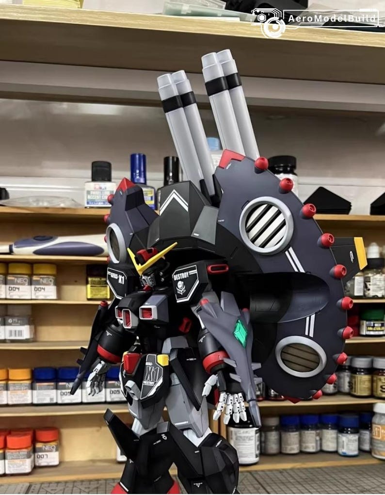 May include: A detailed model of a robot with a black, gray, and red color scheme. The robot has large shoulder-mounted cannons and intricate detailing, including a shield with red accents and a diamond-shaped green emblem. The model is set against a backdrop of shelves with paint bottles.