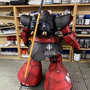 May include: A detailed model of a red and black mecha robot with a yellow spear. The robot has the text "PRINCIPALITY OF ZEON" on its skirt. The model is set against a backdrop of shelves filled with paint bottles and tools.