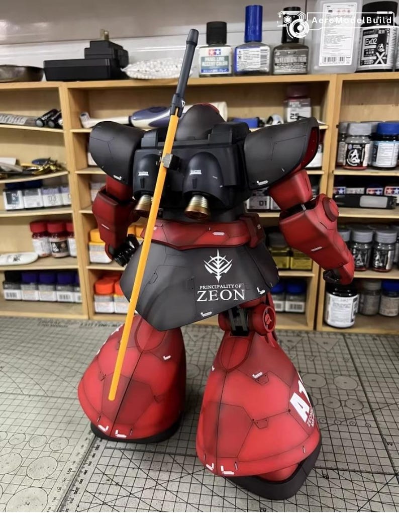 May include: A detailed model of a red and black mecha robot with a yellow spear. The robot has the text "PRINCIPALITY OF ZEON" on its skirt. The model is set against a backdrop of shelves filled with paint bottles and tools.