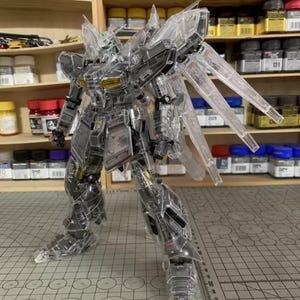 May include: A transparent model robot with articulated joints and wing-like structures. The robot is posed on a cutting mat with a grid pattern. The background includes shelves with paint bottles and model-building supplies. The robot's design features intricate details.