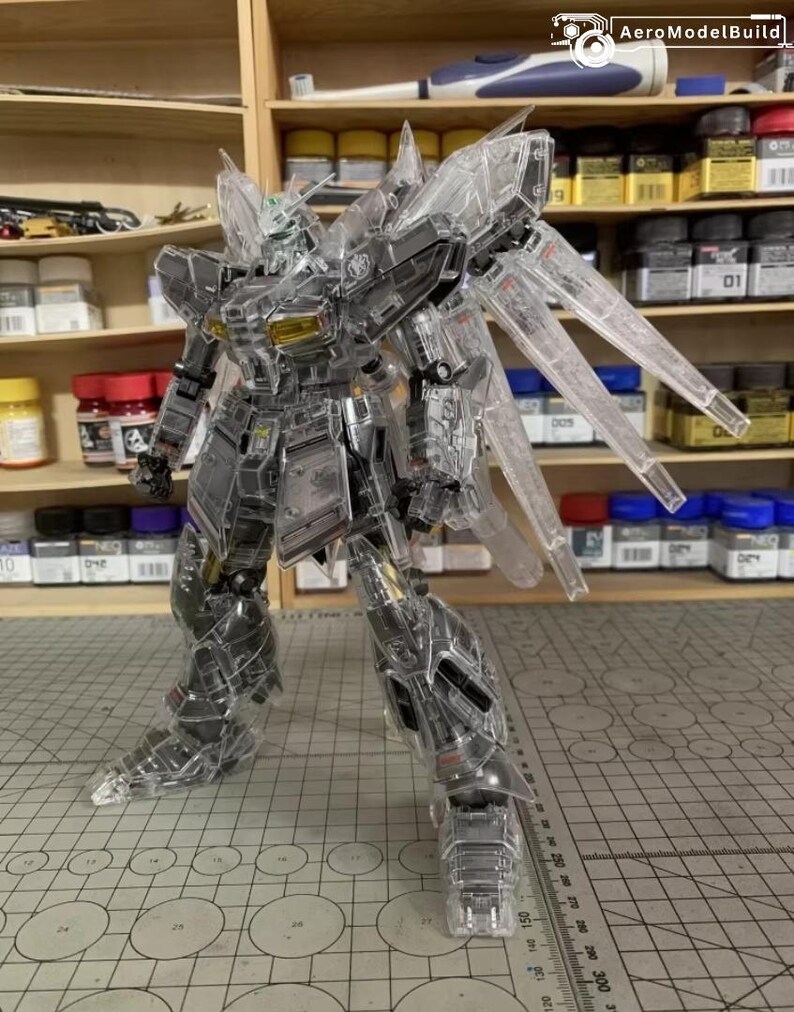 May include: A transparent model robot with articulated joints and wing-like structures. The robot is posed on a cutting mat with a grid pattern. The background includes shelves with paint bottles and model-building supplies. The robot's design features intricate details.