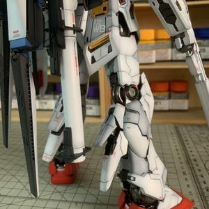 K&ouml;nnte beinhalten: Detaillierter, teilmontierter Modellbausatz eines Mech-Roboters. Das Modell hat wei&szlig;e, graue, rote und schwarze Komponenten. Der Text "102 WB" ist auf dem Bein sichtbar. Das Modell steht auf einer Rasteroberfl&auml;che.