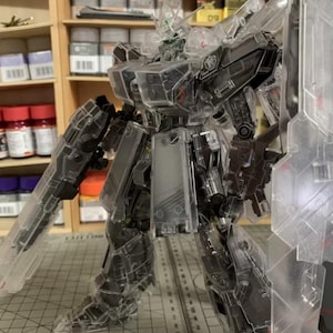 May include: A transparent model robot with visible internal mechanisms. The robot is armed with a gun and a shield. It stands on a cutting mat with metric measurements. Shelves with paint bottles are visible in the background.