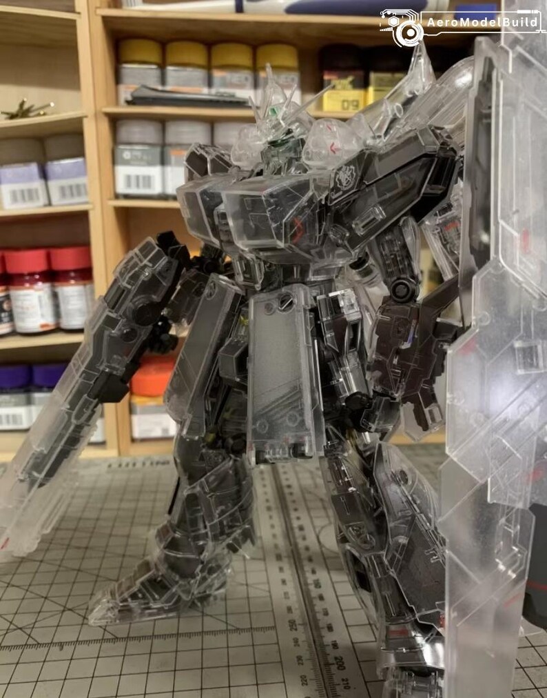 May include: A transparent model robot with visible internal mechanisms. The robot is armed with a gun and a shield. It stands on a cutting mat with metric measurements. Shelves with paint bottles are visible in the background.