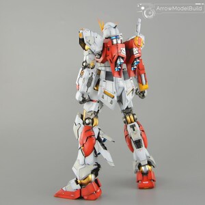 May include: A detailed model robot, predominantly white, red, and gold. The model features intricate mechanical detailing, including joints, panels, and weapons. The robot is posed with one leg forward, highlighting its design. The text "ArrowModelBuild" is visible.